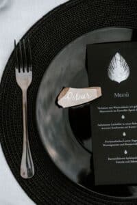 Ein schwarzer Teller mit einer Gabel ruht auf einem gewebten Tischset, perfekt für eine elegante Hochzeit. Ein zerrissenes Stück Papier mit der Aufschrift „Oskar“ wird sorgfältig auf den Teller gelegt, neben einer schwarzen Menükarte mit einem Muschelmuster, das jeder Hochzeitsplanung Charme verleiht.