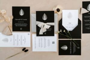Eine flache Lage elegant gestalteter schwarz-weißer Hochzeitseinladungen, die mit einem Blattmotiv die Essenz einer Hochzeit perfekt einfangen. Die Karten sind mit Bändern und zarten Trockenblumen auf einer beigen Oberfläche geschmückt, eine ideale Note für Ihre Hochzeitsplanung.