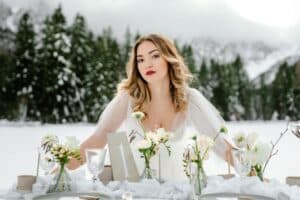 Eine Frau im weißen Kleid steht hinter einem mit Blumen und Zahlenkarten geschmückten Tisch und verkörpert den Zauber der Hochzeitsplanung in einer verschneiten, bewaldeten Landschaft. Die majestätischen Berge im Hintergrund runden diese zauberhafte Kulisse zum Heiraten ab.
