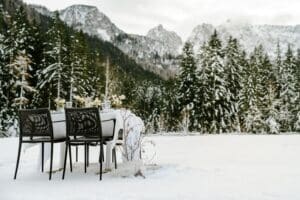 Ein gedeckter Tisch steht auf einem schneebedeckten Feld vor einem Wald, perfekt für eine zauberhafte Hochzeit. Der mit einem weißen Tuch und Blumen geschmückte Tisch fängt die Essenz einer romantischen Hochzeit ein. Schneebedeckte Berge stehen majestätisch im Hintergrund unter einem wolkigen Himmel.