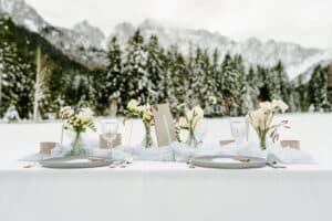 Ein wunderschön gedeckter Esstisch im Freien für eine traumhafte Hochzeit, geschmückt mit weißen Tellern, Kristallgläsern und kleinen Blumenarrangements in einer verschneiten Landschaft. Hohe Bäume und schneebedeckte Berge schaffen eine malerische Kulisse, die sich perfekt für jede Hochzeitsplanung eignet.