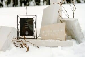 Ein kleines gerahmtes Schild mit deutschem Text und eine Karte mit der Aufschrift „All you need is SWEETS“ liegen eingebettet zwischen weißen rechteckigen Blöcken und Schnee und schaffen die Kulisse für eine romantische Winterhochzeit. Überall sind trockene Zweige und Pflanzen verstreut, perfekte Akzente für Ihre Hochzeitsplanung.