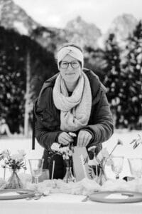 Eine Person in Winterkleidung, mit Brille und Stirnband, arrangiert elegant Blumen auf einem gedeckten Esstisch für eine Hochzeit im Freien. Schneebedeckte Bäume und Berge verstärken die gemütliche, winterliche Atmosphäre und schaffen eine zauberhafte Kulisse, die sich perfekt für die Hochzeitsplanung eignet.