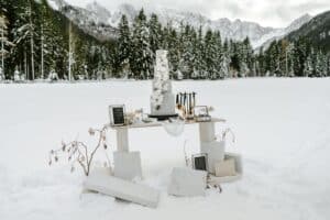 Ein kleiner Tisch in einer verschneiten Landschaft, perfekt für eine Winterhochzeit, enthält verschiedene Gegenstände wie Kerzen, Rahmen und Dekorationsartikel. Schneebedeckte Bäume und Berge in der Ferne umgeben ihn und schaffen eine ruhige Umgebung, die sich ideal für eine romantische Hochzeitsplanung vor einer atemberaubenden Winterkulisse eignet.