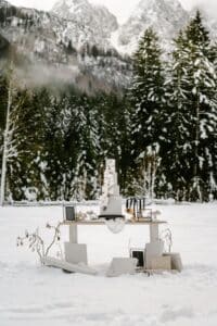 Ein wunderschön gedeckter Hochzeitstisch in einem verschneiten Wald mit einer großen, eleganten weißen Torte. Die Kulisse besteht aus schneebedeckten Bäumen und majestätischen Bergen und schafft eine ruhige Winterlandschaft, die perfekt für diejenigen ist, die ihre Traumhochzeit planen.