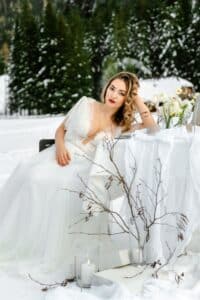 Eine Frau in einem weißen Kleid sitzt an einem schneebedeckten Tisch im Freien, umgeben von winterlichem Grün. Der Tisch, der für eine elegante Hochzeit geeignet ist, ist mit weißen Blumen und Kerzen geschmückt, und davor sind kahle Zweige ausgestellt. Sie blickt mit heiterem Gesichtsausdruck in die Kamera und fängt ihre Hochzeitsplanungsvision ein.
