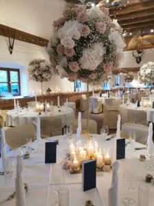 Eleganter Hochzeitsempfang mit runden Tischen, geschmückt mit hohen Blumengestecken mit rosa und weißen Blumen. Perfekt für alle, die schon mitten in der Hochzeitsplanung stecken. Die Tische sind mit weißen Tischdecken, Kerzen, Glaswaren und marineblauen Menükarten gedeckt. Warmes Licht sorgt für eine gemütliche Atmosphäre.