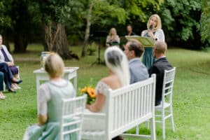 Eine Hochzeitszeremonie im Freien findet statt, bei der eine Frau an einem Podium spricht. Braut und Bräutigam sitzen ihr auf weißen Stühlen gegenüber, während die Gäste in der Nähe sitzen. Bäume und Gras umgeben den Bereich und schaffen eine natürliche Umgebung, die sich perfekt für alle Hochzeitsplanungsbedürfnisse eignet.