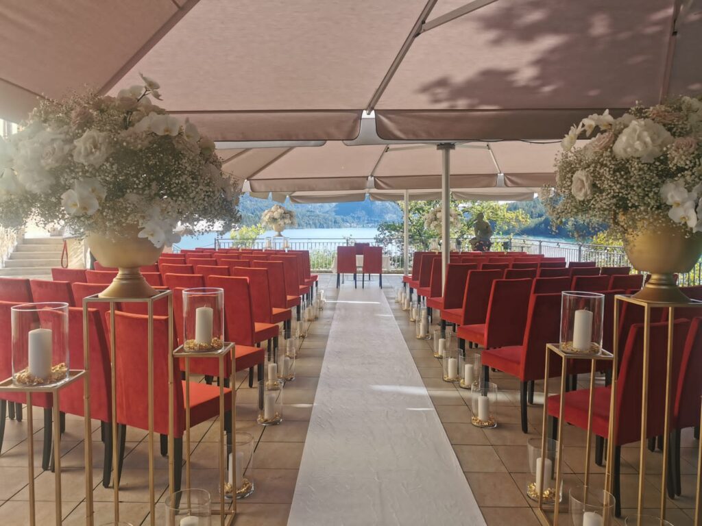 Eine Hochzeitszeremonie im Freien besteht aus Reihen roter Stühle, die auf einen mit Kerzen gesäumten Gang blicken. Große Blumenarrangements mit weißen Blumen stehen auf hohen Ständern. Der Raum, der sich ideal für die Hochzeitsplanung eignet, ist durch Vordächer beschattet und bietet einen malerischen Blick auf Bäume und Wasser.