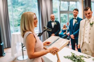Eine Frau in einem rosa Kleid steht an einem Rednerpult mit einem Mikrofon und spricht während der Hochzeitszeremonie. Zwei Bräutigame in formeller Kleidung, einer in Weiß und einer in Blau, stehen ihr gegenüber. Die Gäste sitzen im Hintergrund in der Nähe großer Fenster, alles Teil dieser wunderschön orchestrierten Hochzeitsplanung.