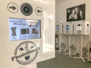 Ein Raum mit mehreren Fotokabinen, perfekt für die Hochzeitsplanung. Im Vordergrund ist eine Touchscreen-Oberfläche auf einer Kabine mit Optionen und einem Kameraobjektiv darüber zu sehen. Im Hintergrund ist an der Wand ein Poster zu sehen, das Leute zeigt, die Spaß haben, vielleicht bei einer Hochzeit.