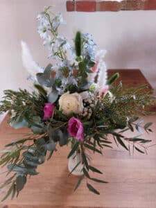 Ein Blumenstrauß in einer Vase auf einem Holztisch, perfekt für die Hochzeitsplanung, mit weißen Rosen, rosa Pfingstrosen und hellblauem Rittersporn. Akzentuiert mit Grünpflanzen wie Eukalyptusblättern und Pampasgras steht er vor einer verschwommenen Backsteinmauer im Hintergrund, ideal für die Romantik einer Hochzeit.