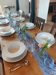 Ein hölzerner Esstisch ist elegant mit weißen Tellern, Schüsseln, Besteck und Weingläsern gedeckt, perfekt für eine Hochzeitsfeier. Kleine Blumenarrangements in Glasgefäßen und ein blauer Stoffläufer schmücken die Mitte dieser anmutigen Einrichtung und schaffen eine bezaubernde Atmosphäre für Ihren besonderen Tag.