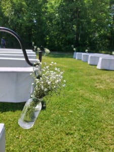 Eine Nahaufnahme weißer Blumen in einem kleinen Glasgefäß hängt an einem Haken und fängt die Essenz einer intimen Hochzeitsplanung ein. Der Hintergrund zeigt ordentlich angeordnete weiße Bänke auf einem grünen Rasen, vor üppigen, belaubten Bäumen unter einem sonnigen Himmel.