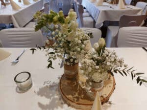 Ein wunderschön gedeckter Tisch mit weißen Tischdecken und eleganten Blumendekorationen in Glasgefäßen ist perfekt für eine anspruchsvolle Hochzeitsplanung. Das Ambiente mit ordentlich gefalteten Servietten und kleinen Kerzen schafft eine einladende Atmosphäre, die sich ideal für jede Hochzeitsfeier eignet.