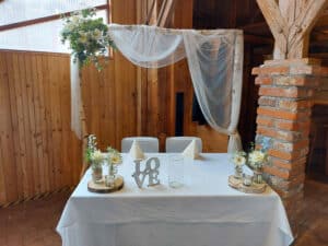 Ein Tisch für den Hochzeitsempfang ist mit einer weißen Tischdecke, Blumenarrangements und einem dekorativen „LOVE“-Schild gedeckt. Dahinter steht ein mit transparentem Stoff und Grünpflanzen behangener Holzbogen in einer rustikalen Umgebung mit Holzwänden und einer Ziegelsäule – eine perfekte Szene für Ihre Hochzeitsplanung.