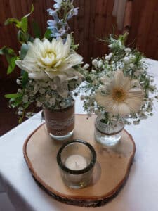 Zwei Glasgefäße mit weißen Blumen, Grünpflanzen und Schleierkraut stehen auf einer Holzscheibe. Vor den Gläsern auf einer weißen Tischdecke leuchtet eine kleine Votivkerze und verleiht dem Ganzen zusätzliche Eleganz. Die rustikale Holzvertäfelung im Hintergrund ist perfekt für die Hochzeitsdekoration und vervollständigt diese charmante Vignette.