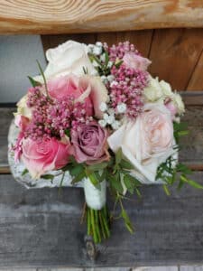 Ein atemberaubender Hochzeitsstrauß mit rosa und weißen Rosen, Schleierkraut und Grünpflanzen, elegant mit einer weißen Schleife zusammengebunden. Er ruht anmutig auf einer verwitterten Holzoberfläche und ist perfekt für Ihre Hochzeitsplanung.