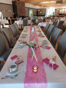 Ein langer Esstisch ist elegant für eine Hochzeitsplanungsveranstaltung gedeckt, mit einem rosa Tischläufer, weißem Geschirr, rosa gefalteten Servietten und kleinen Blumendekorationen. Der Raum ist mit grauen Stühlen und warmen Holzakzenten ausgestattet, mit einem Dekor, das „Liebe“ an die Wand schreibt.