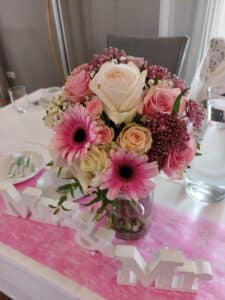 Ein Strauß rosa Rosen, Gerbera und weißer Blumen ziert eine klare Vase auf einem Tisch mit rosa Tischläufer. Im Vordergrund lugt ein „Mr & Mrs“-Schild hervor und deutet auf das freudige Ereignis der Hochzeit hin. Gläser und Geschirr bilden einen eleganten Hintergrund für diese intime Hochzeitsplanungsszene.