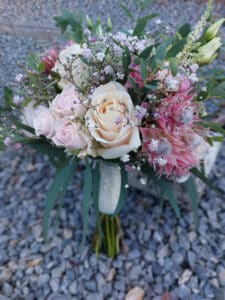 Ein perfekter Hochzeitsstrauß mit blassrosa und cremefarbenen Rosen, akzentuiert mit kleinen weißen Blüten und rosa Proteas, umgeben von üppigem Grün. Dieses elegante Arrangement sieht auf einer Kiesfläche wunderschön aus.