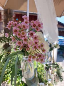 An einer Schnur hängt eine kleine Glasflasche, gefüllt mit Wasser und einem Strauß zartrosa Wachsblumen und Grünpflanzen. Eine weiße Rose schmiegt sich zwischen die Blumen und ist perfekt für jede Hochzeit. Der Hintergrund zeigt eine sonnige Außenkulisse mit verschwommenem Grün und rustikalen Strukturen.