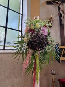 Ein üppiges Blumenarrangement mit Rosen, Hortensien und Grünpflanzen auf einem Holzständer schmückt den Innenraum der Kirche und ist perfekt für eine Hochzeit. Buntglasfenster und ein Kruzifix sorgen für eine ruhige Kulisse und machen die Kirche zu einem idealen Ort für die Hochzeitsplanung.