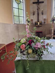 Ein Blumenarrangement mit rosa, violetten und weißen Blumen schmückt einen mit grünem Tuch bedeckten Tisch in der Kirche, perfekt für jede Hochzeit. Buntglasfenster und ein Kreuz bestimmen die Szene im Hintergrund.