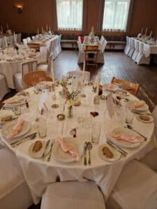 Ein wunderschön dekorierter Bankettsaal, ideal für eine Hochzeit, mit elegant gedeckten runden Tischen. Jeder Tisch ist mit weißen Tischdecken, gefalteten Servietten, Kerzen und Blumenschmuck ausgestattet. Die Sitzplätze sind mit Holzstühlen ausgestattet, während natürliches Licht durch die großen Fenster fällt, perfekt für Ihre Hochzeitsplanung.