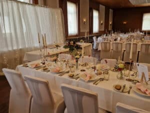 Eleganter Hochzeitsempfangssaal mit dekorierten Tischen mit weißen Tischdecken, rosa Servietten, Blumengestecken und brennenden Kerzen. Perfekt für Ihre Hochzeit, schmücken Lichterketten den Hintergrund und schaffen eine warme und einladende Atmosphäre, ideal für diejenigen, die gerade mitten in der Hochzeitsplanung sind.