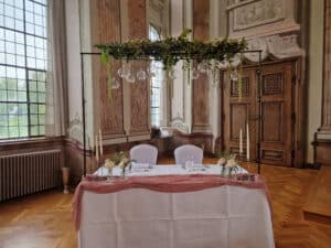 Ein wunderschön dekorierter Hochzeitssaal im Innenbereich mit zwei Stühlen an einem mit einem rosa Tuch geschmückten Tisch, Blumenarrangements, Kerzen und hängenden Glaskugeln. Der Raum verfügt über kunstvolle Holztüren, große Fenster und klassische architektonische Details, die perfekt für Ihre Traumhochzeitsfeier sind.