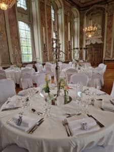 Ein luxuriöser Bankettsaal, der sich perfekt für Hochzeitsfeiern eignet. Er bietet elegant gedeckte, runde Tische, die mit weißen Tischdecken und Stuhlhussen geschmückt sind. Jeder Tisch verfügt über einen hohen Kerzenleuchter als Tafelaufsatz, edles Glasgeschirr und ordentlich angeordnetes Besteck, alles unter kunstvollen Kronleuchtern und aufwendigen architektonischen Details.