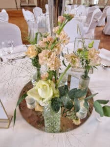 Ein Blumengesteck mit hellrosa und cremefarbenen Blumen ziert den Tisch, arrangiert in Glasvasen auf einem verspiegelten Sockel. Der elegant gedeckte Tisch mit weißen Tischdecken und Stühlen ist perfekt für eine anspruchsvolle Hochzeit und wertet Ihr Heiratserlebnis auf.