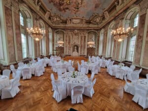 Ein großer Speisesaal für eine Hochzeitsfeier mit runden, mit weißen Tischdecken bedeckten Tischen, die mit Tafelaufsätzen geschmückt und von eleganten Stühlen umgeben sind. Verzierte Kronleuchter hängen von einem detailreichen Deckengemälde herab und der Raum bietet hohe Fenster und eine üppige Dekoration, die perfekt für die Hochzeitsplanung ist.