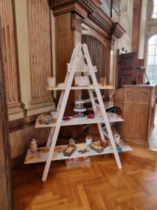 Eine weiße, leiterartige Auslage präsentiert verschiedene Snacks und Süßigkeiten in Glasbehältern in einem eleganten Raum mit Holzvertäfelung und großen Fenstern – perfekt für jede Hochzeitsfeier.