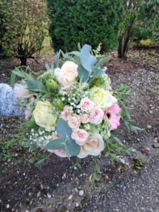 Ein Strauß blassrosa Rosen, weißem Schleierkraut und Grünpflanzen, darunter Eukalyptusblätter, perfekt für eine Hochzeit, wird vor einer Gartenkulisse mit Kies und Sträuchern präsentiert.