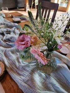 Ein Tisch mit blaugrünem Stoff ist mit zwei Glasvasen geschmückt, in denen rosa, weiße und grüne Blumen stehen. Das Arrangement, perfekt für die Hochzeitsplanung, umfasst Gänseblümchen, Lisianthus und Grünpflanzen, um ein frisches und elegantes Herzstück für Ihre Hochzeit zu schaffen.