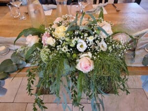 Ein Blumenarrangement mit rosa und weißen Rosen, umgeben von üppigem Grün und zarten weißen Blüten, ziert den mit Gläsern und Tellern gedeckten hölzernen Esstisch und verleiht Ihrer Hochzeitsplanung einen Hauch von Eleganz.
