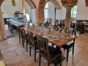Ein gemütliches Restaurant mit Holztischen zum Essen, perfekt für alle, die eine Hochzeit planen. Jeder Tisch ist mit Grünpflanzen, Kerzen und ordentlich angeordnetem Besteck dekoriert. Gewölbte Ziegeltüren und große Fenster verleihen dem Raum einen rustikalen Charme.