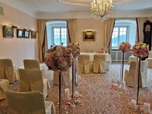 Ein kleiner Trauungsraum mit eleganter Dekoration, perfekt für Ihre Hochzeit. Die Stühle sind mit beigem Stoff bezogen und Blumenarrangements mit rosa und weißen Blüten säumen den Gang. Ein Kronleuchter hängt von der Decke und große Fenster sorgen für natürliches Licht bei Ihrer Hochzeit.