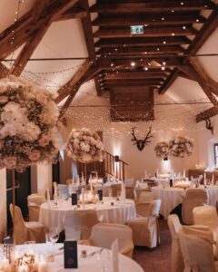 Bei einem rustikalen Hochzeitsempfang stehen runde Tische mit weißen Tüchern und Stühlen mit cremefarbenen Hussen. Blumengestecke und warme Lichterketten schmücken die Holzbalken und sorgen für eine gemütliche Atmosphäre. Kerzen sorgen für sanfte Beleuchtung und Geweihe schmücken die Wand.