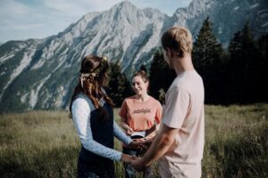 Ein Paar hält sich auf einer Bergwiese an den Händen, während es einer Frau gegenübersteht, die die Trauung durchzuführen scheint. Im Hintergrund sind eine Reihe majestätischer Berge und Kiefern unter einem klaren blauen Himmel zu sehen.