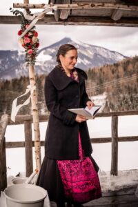 Eine Person in schwarzem Mantel und rosa Kleid steht auf einem verschneiten Balkon und hält ein Buch in der Hand. Dahinter ist eine Berglandschaft unter einem wolkigen Himmel zu sehen – eine perfekte Kulisse für Hochzeitsplanung. Ein Birkenbogen mit Blumen und Bändern verleiht Charme, während im Vordergrund ein weißer Eimer zu sehen ist.