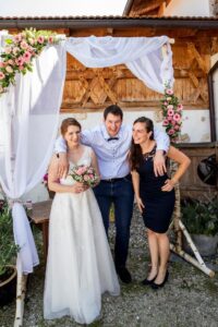 Eine Braut in einem weißen Kleid hält einen Blumenstrauß und lächelt neben einem Mann in einem weißen Hemd und einer Frau in einem schwarzen Kleid, die unter einem mit rosa und weißen Blumen geschmückten Torbogen stehen. Diese malerische Hochzeitsszene spielt sich draußen vor dem Hintergrund einer charmanten Holzkonstruktion ab.