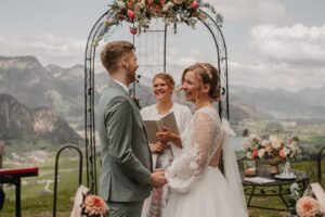 Ein Paar gibt sich in einer wunderschön geplanten Hochzeitszeremonie im Freien freudig das Ja-Wort. Sie stehen unter einem Blumenbogen mit einer malerischen Bergkulisse im Hintergrund. Der Bräutigam trägt einen Anzug und die Braut ein Spitzenkleid. Zwischen ihnen steht ein lächelnder Geistlicher, der ein Buch in der Hand hält.