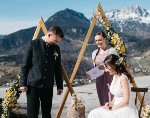 Ein Paar heiratet im Freien vor einer malerischen Bergkulisse und verkörpert eine exquisite Hochzeit. Die Braut sitzt in einem weißen Kleid, der Bräutigam steht in einem dunklen Anzug. Eine Frau amtiert die Trauung und hält ein Notizbuch in der Hand. Blumendekorationen und Holzkonstruktionen rahmen die fachmännisch geplante Szene ein.