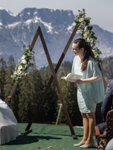 Eine Frau steht mit einem Buch in der Hand auf einer grünen Plattform vor einem dreieckigen Holzbogen, der mit weißen Blumen und Grün geschmückt ist – perfekt für jede Hochzeit. Schneebedeckte Berge und Bäume bilden die idyllische Kulisse für diese traumhafte Hochzeitskulisse.