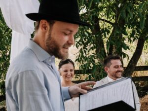 Eine Person mit Hut liest aus einem Buch, während ein lächelndes Paar in der Nähe unter einem Baum sitzt und vielleicht über seine bevorstehende Hochzeit spricht. Die Szene wirkt entspannt und sonnig, mit viel Grün im Hintergrund.