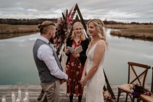 Ein Paar steht vor einem dreieckigen Blumenbogen am See und bereitet sich auf seine Hochzeit vor. Die Braut trägt ein weißes Kleid und der Bräutigam Weste und Krawatte. Ein bewölkter Himmel wirft ein sanftes Licht auf ihre Hochzeit, während sie dem lächelnden Geistlichen gegenüberstehen und das ruhige Wasser im Hintergrund sehen.