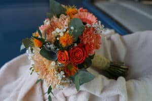 Ein lebendiger Blumenstrauß mit orangefarbenen Rosen, rosa Gerbera und zartpfirsichfarbenen Dahlien, akzentuiert mit weißem Schleierkraut und grünen Blättern, ruht anmutig auf einem cremefarbenen Stoff – perfekt für diese besonderen Momente bei der Hochzeitsplanung.